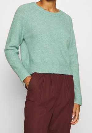 Personne portant un pull pelucheux vert clair et un pantalon bordeaux taille haute, une main dans la poche, sur un fond uni.