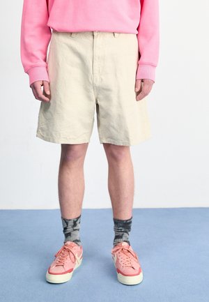 Osoba ubrana w beżowe szorty, różową bluzę, czarno-szare skarpetki tie-dye oraz różowe sneakersy Nike stojąca na niebieskim dywanie.