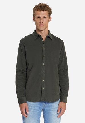 Mann mit kurzen lockigen Haaren, der ein dunkelgrünes, langärmeliges Hemd mit Knöpfen und hellblaue Jeans trägt, steht vor einem schlichten Hintergrund.