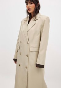 Beige double-breasted coat with gold buttons, stitched lapels, two front pockets, and brown cuffs peeking from underneath the sleeves.