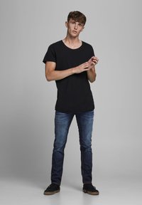 Joven de pie contra un fondo gris, vestido con una camiseta negra, jeans azules y zapatillas negras, con las manos entrelazadas en movimiento.