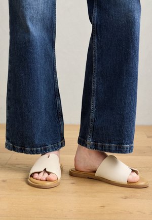Fødder iført beige slidesandaler med udskæringsdetaljer, kombineret med mørkeblå vidde jeans, stående på trægulv.