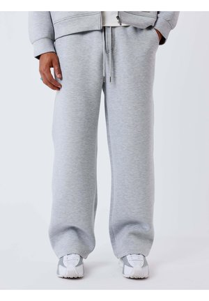 Bas du corps portant un pantalon de survêtement gris clair avec cordon de serrage et des baskets grises assorties sur un fond blanc uni.