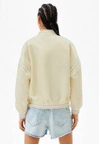 Jasno beżowa kurtka bomberka z elastycznym pasem i mankietami, z dwoma pionowymi paskami na rękawach. Nosi ją na spodenkach dżinsowych.