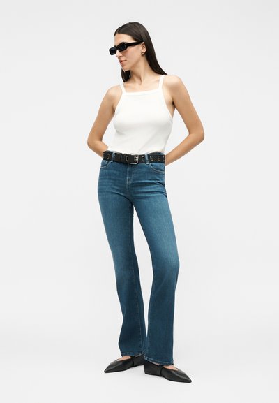 Γυναίκα με μαύρα γυαλιά ηλίου, λευκό αμάνικο τοπ, μπλε τζιν ψηλόμεσο με μαύρη ζώνη και μαύρες μυτερές μπαλαρίνες, που στέκεται με τα χέρια πίσω από την πλάτη.