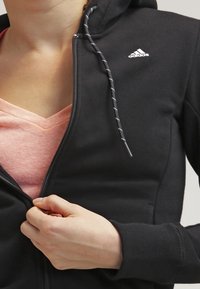 Hoodie zippé noir avec une finition texturée, présentant une capuche avec cordon de serrage et un logo blanc sur la poitrine gauche. Superposé sur une chemise rose.