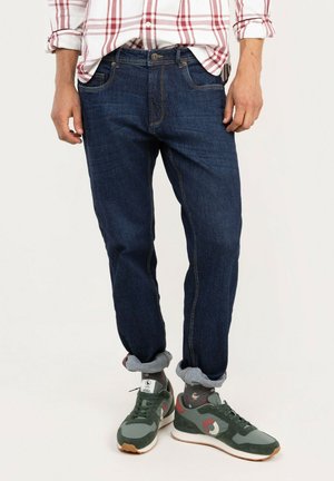 Hombre que lleva vaqueros azul oscuro remangados, calcetines grises con estampado, zapatillas verdes y grises, y una camisa blanca con rayas rojas.