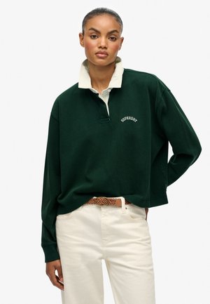 Polo-shirt à manches longues vert foncé avec un col crème, comprenant une patte de boutonnage à l'avant et un subtil logo brodé "Superdry". Associé à un pantalon crème.