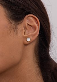 Goldfarbene Steckohrringe mit einer runden, glatten weißen Oberfläche. Im Vordergrund eine Nahaufnahme des Ohrs eines Modells, die das Design des Ohrrings hervorhebt.