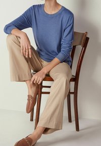 Pull en maille bleu à col rond, associé à un pantalon beige à jambes larges et des mocassins marron ornés d'un accent chaîne. Assis sur une chaise en bois.