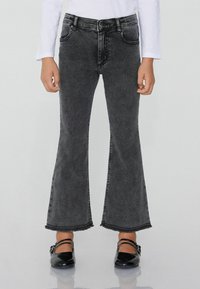 Dítě v černých zkrácených flare džínách s třepicími lemy, bílé dlouhé tričko a černé lesklé Mary Jane boty.