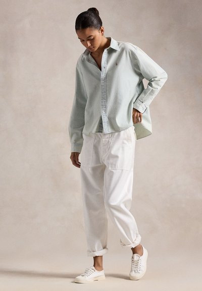 Sötét hajú nő kontyba fogva világoszöld, túlméretezett, gombos inget visel, fehér, lazán szabott nadrágot hajtott szárakkal és fehér sportcipőt.