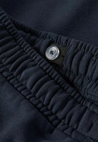 Granatowa tkanina z elastycznym pasem i przezroczystym plastikowym guzikiem. Charakteryzuje się marszczoną teksturą i detalem przeszyć, podkreślającym jakość materiału.