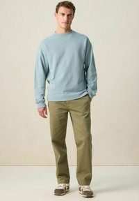 Jeune homme avec des cheveux bouclés, portant un sweat-shirt bleu clair, un pantalon olive et des baskets grises, se tenant devant un fond beige uni.