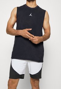 Maglietta atletica senza maniche nera con logo bianco, abbinata a pantaloni corti bianchi con dettagli e inserti neri. Progettata per lo sport.
