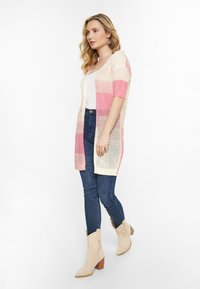 Cardigan lungo fino al ginocchio in maglia aperta con strisce alternate rosa e crema, indossato sopra una maglietta bianca con jeans in denim blu e stivali alla caviglia beige.
