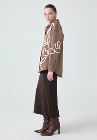 Brun oversized skjorta med vit blommig broderi, kombinerad med en lång brun kjol och bruna högklackade stövlar. Slät textur, avslappnad passform.