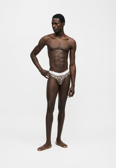 Λεπτός, σκουρόχρωμος άνδρας στέκεται ξυπόλητος, φοράει εσώρουχα με λεοπάρ σχέδιο και λευκή ζώνη, κοιτάζει αριστερά σε απλό γκρίζο φόντο.