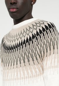 Strickpullover mit Rundhalsausschnitt, cremefarbener Basis und geometrischen Mustern in Schwarz und Grün. Strukturierter Stoff mit lockerer Passform.