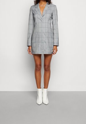 Femme portant une robe blazer à carreaux gris à manches longues et des bottines blanches, debout devant un fond clair uni.