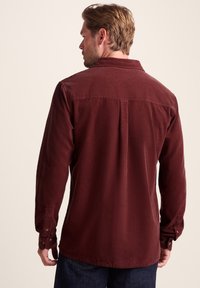 Burgundy corduroy shirt with a button-down collar, long sleeves, and a center back pleat; features button cuffs and a soft texture.