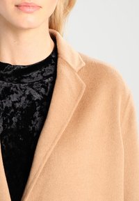 Beige uldfrakke med krave, båret over en sort fløjlsbluse. Frakken har en glat tekstur og en skræddersyet pasform.