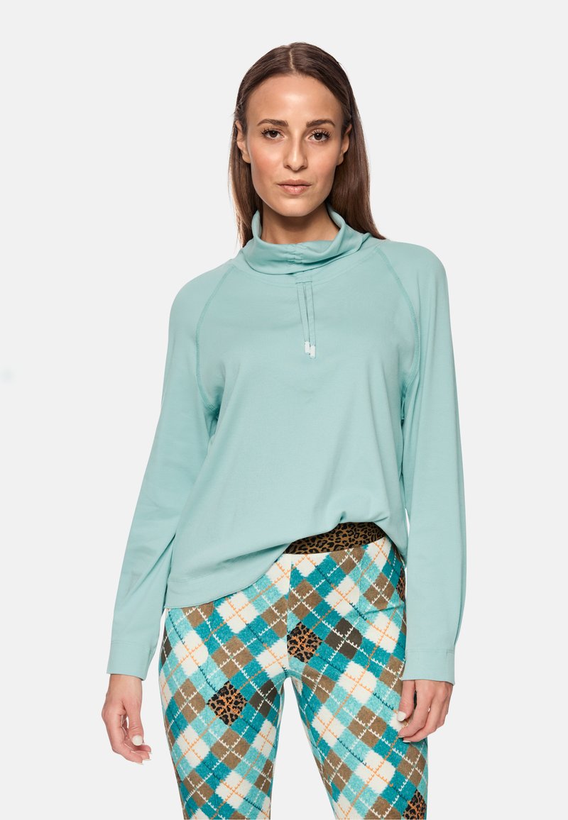 Light turquoise long-sleeve top with a high collar, featuring a half-zip. Paired with patterned leggings in turquoise, brown, and cream.