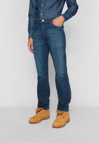 Jeans en denim bleu foncé avec une coupe droite, un design à cinq poches et des détails de couture visibles, associés à des bottes en suède tan.