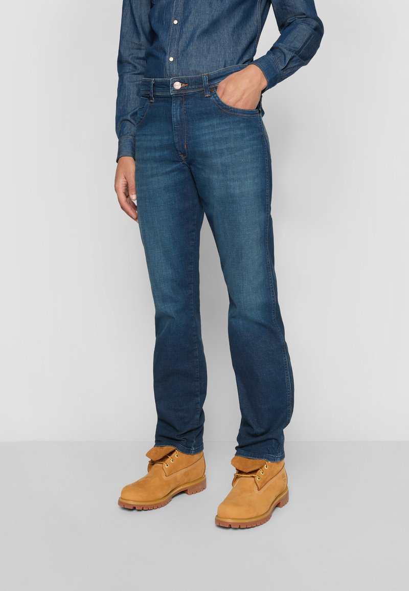 Jeans en denim bleu foncé avec une coupe droite, un design à cinq poches et des détails de couture visibles, associés à des bottes en suède tan.