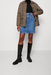 Person som bär en brun tweedjacka, svart polotröja, blå jeanskjol och svarta stövlar i knähöjd mot en enfärgad bakgrund.