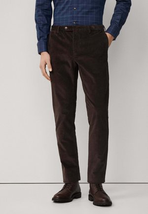 Dunkelbraune Cordhose mit geradem Bein, ausgestattet mit einem Knopfverschluss und Gürtelschlaufen, kombiniert mit braunen Schnürstiefeln.