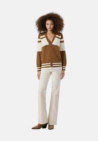 Cardigan a righe marroni e crema con scollo a V e chiusura con bottoni, tessuto a coste, abbinato a pantaloni a campana crema e stivali beige con tacco.