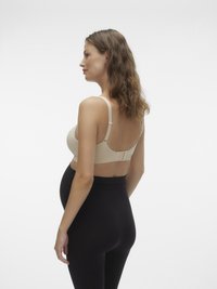 Grūtniece bežā krūšturī un melnās grūtniecības legingās stāv profila skatā uz vienkārša balta fona.