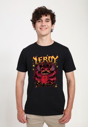 Giovane uomo con capelli ricci sorridente, che indossa una maglietta nera con un mostro dei cartoni animati rosso e il testo "Leroy Deviant Experiment" su sfondo bianco.
