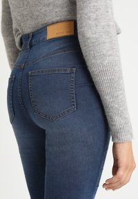 Jeans in denim blu con vita media, dotati di un'etichetta in pelle marrone sulla cintura e due tasche posteriori con un design di cucitura classico.