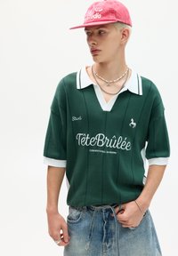 Grøn strikket polo skjorte med hvid krave og horisontale striber, der har "Tête Brûlée" logoet og et lille hesteemblem. Båret med denimshorts.
