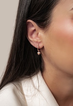 Nahaufnahme eines Frauenohrs mit glattem, dunklem Haar, das einen kleinen goldenen Creolen-Ohrring mit einem herzförmigen Anhänger trägt.