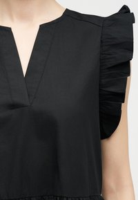 Μαύρο αμάνικο τοπ με βαθύ V σε λαιμόκοψη και λεπτομέρεια με βολάν στους ώμους σε χέρι με ανοιχτό τόνο δέρματος.