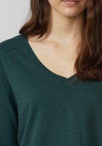 Dunkelgrünes, langärmliges Oberteil mit V-Ausschnitt, aus weichem, strukturiertem Stoff gefertigt. Verfügt über dezente Nähte am Ausschnitt.