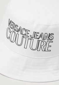 Biely klobúk z textúrovaného materiálu s čiernym potlačeným textom "VERSACE JEANS COUTURE." Dizajn obsahuje zaoblenú korunu a široký okraj.