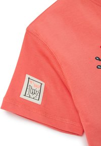 Camiseta de algodón color coral con un diseño gráfico que presenta un gesto de mano, etiqueta con "¡SÍ HERMANO!" en negro sobre un fondo blanco, textura suave.