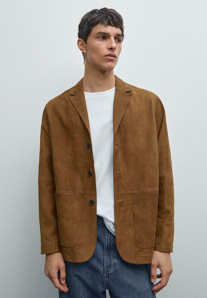Brauner Wildlederblazer mit lockerer Passform, ausgestattet mit einem Reverskragen, zwei Fronttaschen und schwarzem Knopfverschluss. Trägt man über einem weißen T-Shirt.