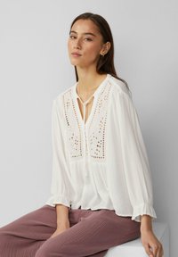 Weißes Blusenshirt aus leichtem Stoff mit Spitzenverzierung, durchgehender Knopfleiste, gebundenem Kragen und Puffärmeln; kombiniert mit weichen, strukturierten rosa Hosen.