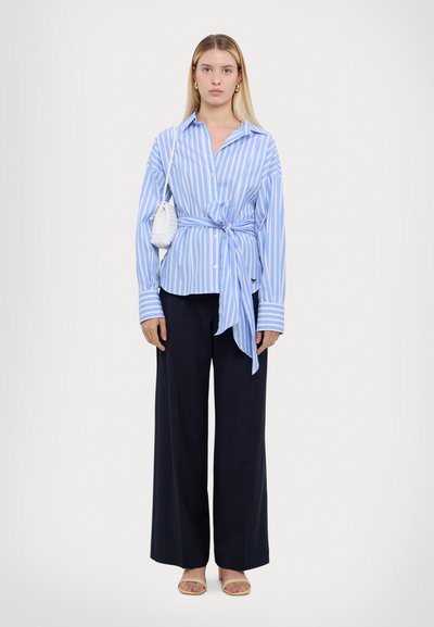 Blauw-wit gestreept overhemd met knoopsluiting en een gestrikte taille, gecombineerd met wijde marineblauwe broeken en gele open teen sandalen. Kleine witte handtas.