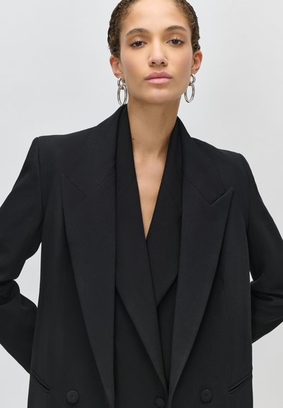 Žena s kudrnatými vlasy v černém oversized saku a stříbrných náušnicích s dvojitými kruhy, hledící přímo vpřed na jednoduchém světlém pozadí.