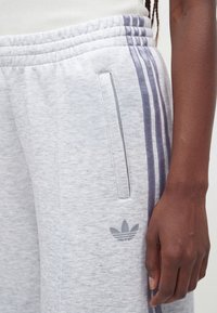 Pantalon de survêtement gris clair avec trois bandes gris foncé sur le côté, poche zippée, logo Adidas proche de la poche, et un bras à la peau foncée à côté.