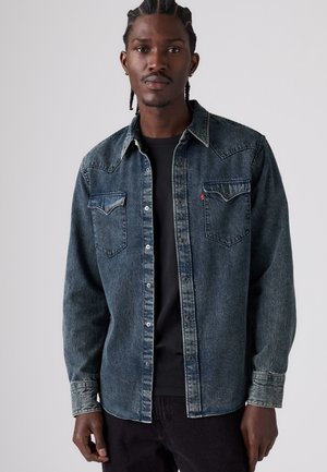 Mann mit geflochtenem Haar, der ein dunkles Denimhemd über einem schwarzen T-Shirt trägt und vor einem schlichten Hintergrund steht.