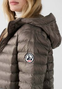 Giacca imbottita kaki con cappuccio, caratterizzata da un design trapuntato, una texture liscia e una toppa circolare sulla spalla sinistra.