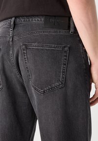 Jean en denim noir avec une coupe classique, deux poches arrière et un patch logo Calvin Klein à la taille. La texture est lisse avec un léger délavage.