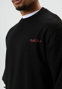 Sweat-shirt noir avec col rond, manches courtes et texte brodé en rouge "Live with Passion Garment Production." Texture de tissu lisse.